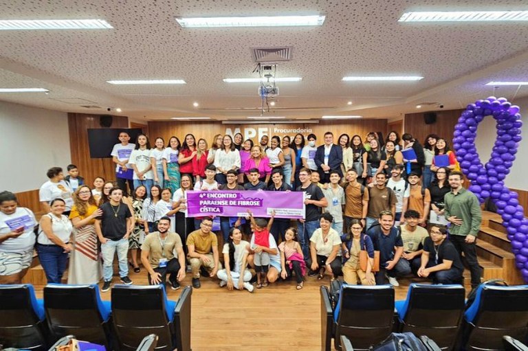 Foto de um grande grupo de pessoas em auditório, de pé e agachadas, segurando uma faixa roxa com os dizeres "4º Encontro Paraense de Fibrose Cística".