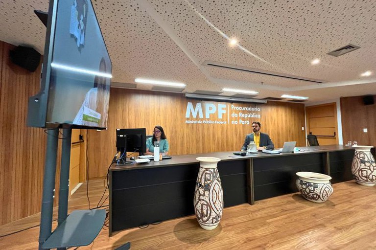 Foto de auditório com duas pessoas em bancada sob painel do MPF Pará, vasos marajoaras e TV em primeiro plano.