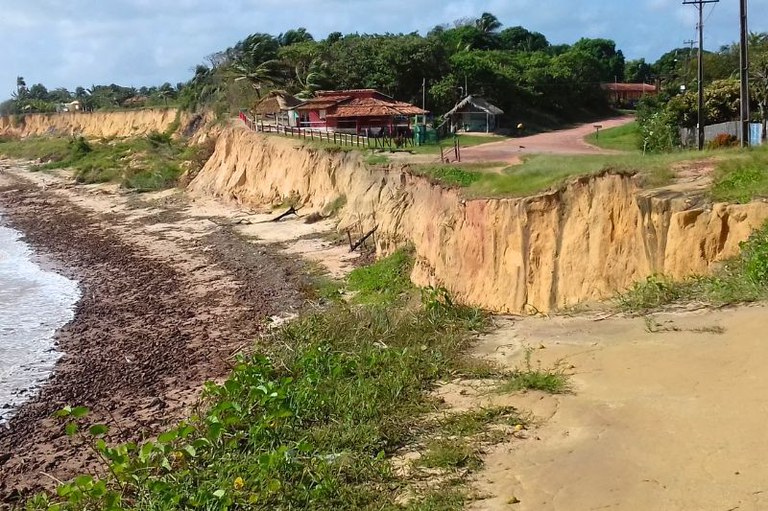 Foto de uma área de erosão costeira em Maracanã, Pará, com falésias de terra amarelada, vegetação e construções no topo, e mar na esquerda.