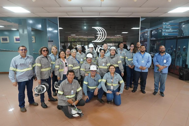 Foto de grupo de pessoas vestidas com uniformes azuis de trabalho, muitos usando capacetes de segurança, posando em um ambiente interno. Eles estão em frente a uma parede com a logo da empresa Hydro.