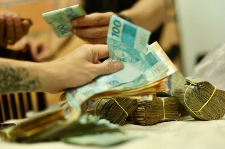 Mãos de pessoas manuseiam notas de R$ 100 ao lado de maços de dinheiro presos por elásticos amarelos sobre uma superfície clara.