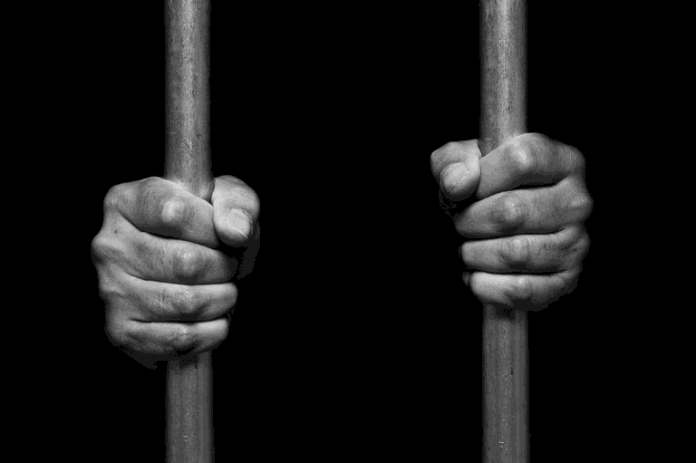 #ParaTodosVerem: A imagem em preto e branco mostra duas mãos segurando barras de metal verticais de uma cela de prisão contra um fundo totalmente preto.