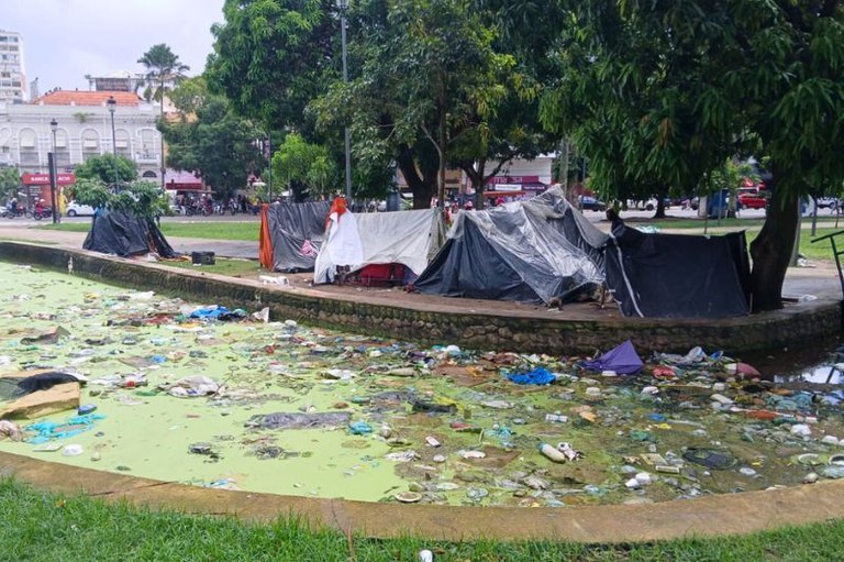 Praça com barracas de lona sob árvores e um canal de água esverdeada coberto por diversos resíduos descartados no primeiro plano.
