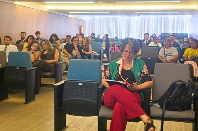 Mulher de calça vermelha e colete verde escreve em um auditório com várias pessoas sentadas nas poltronas ao fundo