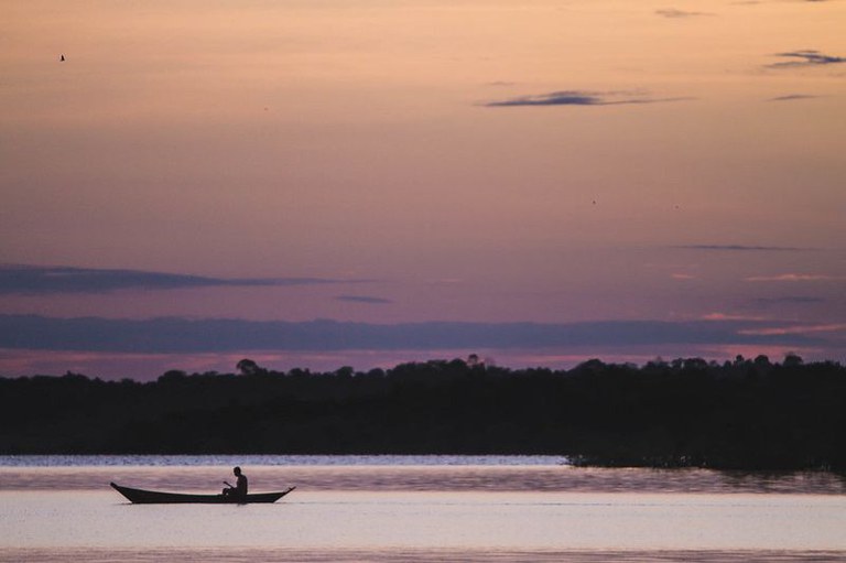 Silhueta de um barco com uma pessoa remando em um rio ao pôr do sol, com o céu em tons de roxo e laranja e uma faixa escura de floresta ao fundo