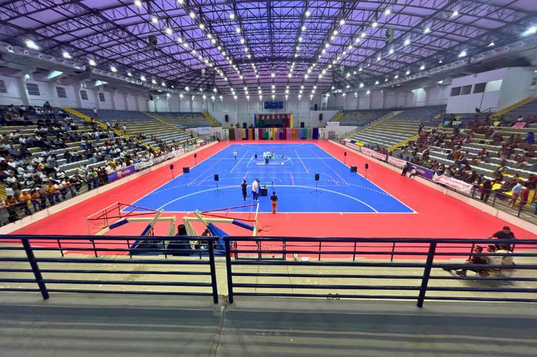 Ginásio amplo com quadra azul e bordas vermelhas, arquibancadas com público e teto metálico iluminado. No centro da quadra, poucas pessoas em pé cercam uma mesa, com equipamentos de som e câmeras distribuídos ao redor da área de jogo.