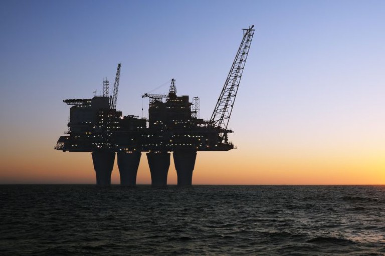 Silhueta de uma plataforma de petróleo iluminada sobre o mar escuro, sob céu azul que se torna alaranjado no horizonte.