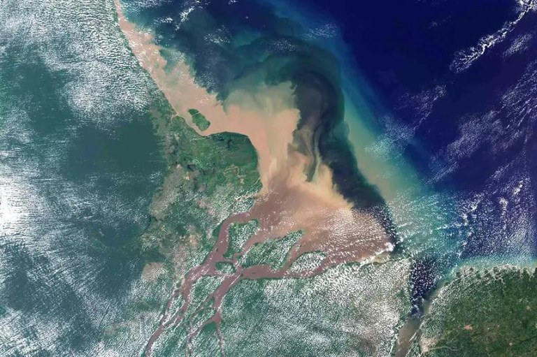 Foto de satélite mostra a foz do rio Amazonas. A pluma marrom do rio desagua no Oceano Atlântico, azul-escuro. A terra ao redor é verde, com algumas nuvens brancas.