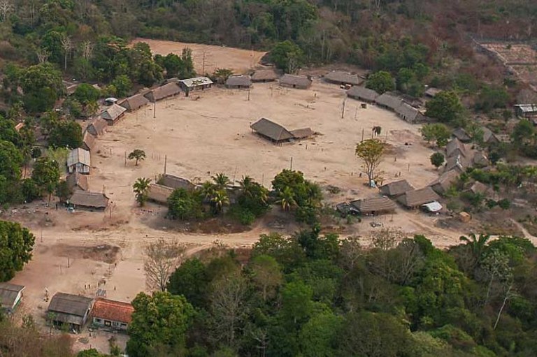 Vista aérea da Terra Indígena Baú, com uma aldeia circular com casas de palha ao redor de um grande pátio central de terra batida, cercada por mata.