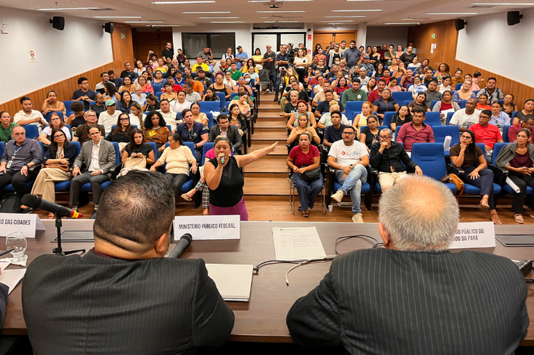 Vista de trás de uma mesa com placas indicando Ministério Público Federal e Ministério Público do Estado do Pará. Uma mulher fala ao microfone diante de um auditório lotado com cadeiras azuis.