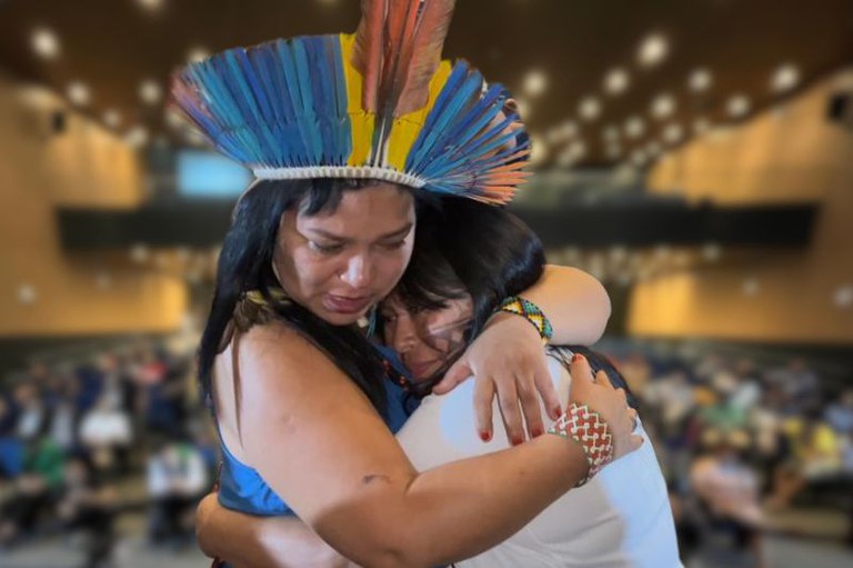 Foto das lideranças indígenas Auricélia Arapiun e Alessandra Korap se abraçando no auditório da Seduc, em Belém, logo após a realização da audiência judicial com diálogo interétnico e intercultural, em 31/01/2025. O fundo desfocado revela várias pessoas sentadas.