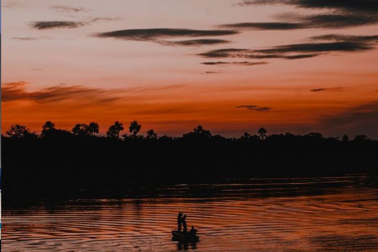 Foto de um pôr do sol com tons de laranja e rosa no céu, refletidos em um rio calmo. Silhuetas de palmeiras e vegetação aparecem ao fundo, enquanto duas figuras estão em um pequeno barco no centro da imagem.