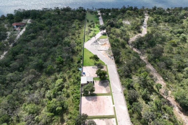 Foto aérea mostra uma área de floresta, ao redor de um caminho pavimentado e várias estruturas. Há divisões de terrenos com vegetação esparsa e algumas construções ao longo do caminho. À direita da imagem, há uma trilha de terra que atravessa a vegetação, e ao fundo, o rio Tapajós.