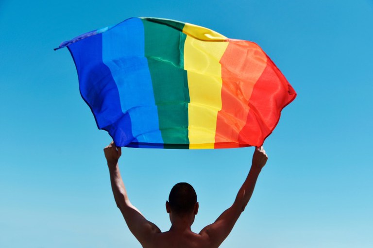 Foto de costas de uma pessoa sem camisa erguendo uma bandeira arco-íris desfraldada ao vento, contra um céu azul sem nuvens