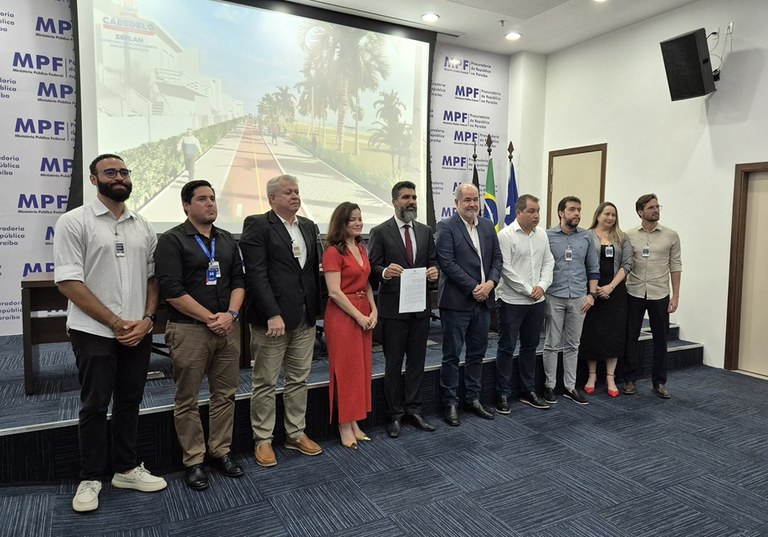 Em uma sala de conferência, pessoas estão alinhadas em pé, lado a lado, em frente a um telão que exibe a imagem renderizada de um projeto urbanístico com palmeiras e calçadas largas. Ao fundo, há uma parede com o logotipo repetido do Ministério Público Federal. Um dos participantes, ao centro, segura um documento.