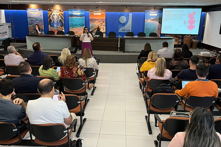 A imagem mostra uma reunião sendo realizada em um auditório. Uma mulher está em pé, falando ao microfone, diante de um público sentado.