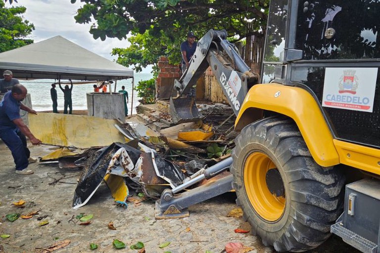 Foto mostra demolição de uma construção irregular na orla, com uma escavadeira e homens recolhendo entulhos.