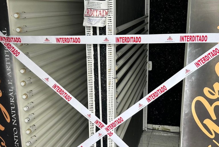 A imagem mostra a entrada de um estabelecimento comercial interditado. Faixas brancas com a palavra INTERDITADO em letras vermelhas estão cruzadas em formato de X na porta, indicando que o acesso está proibido. Acima das faixas, há um aviso impresso reforçando a interdição. Dentro do local, é possível ver painéis verticais com varias lâmpadas fluorescentes.
