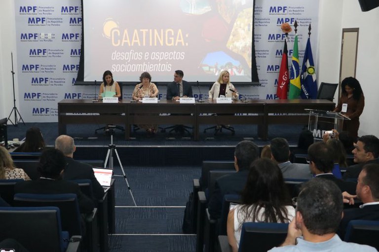 Imagem de mesa retangular com cinco autoridades e plano de fundo com arte do evento da Caatinga