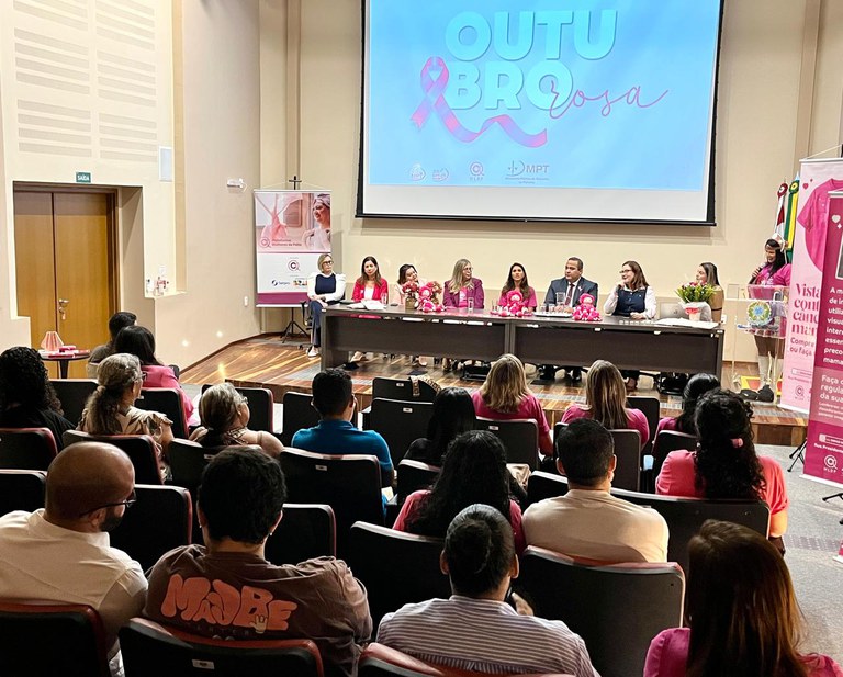 A foto mostra um evento em uma sala de auditório com o tema "Outubro Rosa", dedicado à conscientização do câncer de mama. Na frente, há uma mesa comprida onde estão sentados oito palestrantes, a maioria mulheres, vestidas em tons de rosa ou branco. A plateia é composta por muitas pessoas, algumas também vestindo rosa, sentadas nas poltronas escuras.