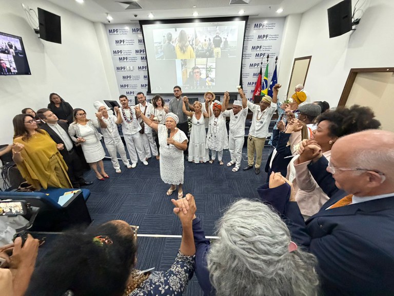 Auditório do MPF com dezenas de pessoas reunidas em círculo, de mãos dadas. No centro, uma mulher vestida de branco fala ao microfone, enquanto integrantes de religiões de matriz africana, também de branco e com guias e adornos tradicionais, participam do momento. Ao fundo, há uma tela de videoconferência, bandeiras do Brasil e do MPF e a identidade visual da instituição nas paredes. O ambiente transmite união, diálogo e respeito entre os participantes.