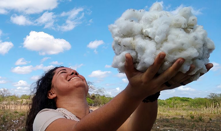 Uma mulher está ao ar livre, sorrindo e olhando para cima enquanto segura nas mãos uma grande porção de algodão branco e fofo.