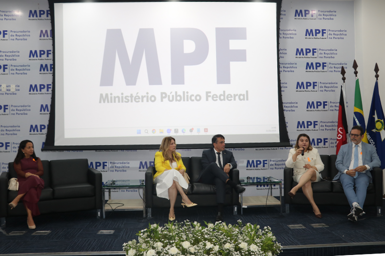 Cinco palestrantes sentados em sofás pretos durante um evento em um palco, posicionados à frente de um grande telão com o logotipo do Ministério Público Federal (MPF), ladeado por bandeiras oficiais à direita.