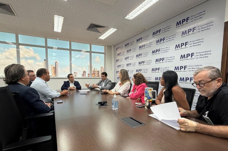 Foto de uma mesa com nove pessoas sentadas ao redor em uma reunião, ao fundo janela com vista da cidade e na parede ao lado, banner com a marca do MPF