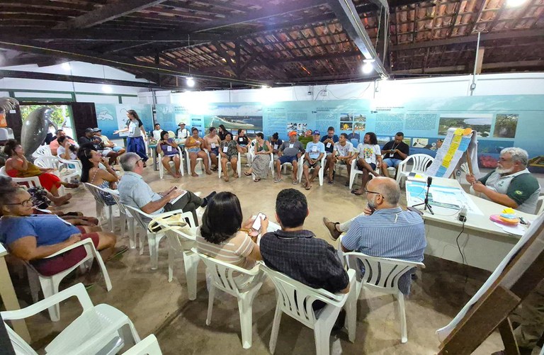 A imagem mostra uma reunião comunitária no Centro de Visitantes da APA da Barra do Rio Mamanguape, um espaço coberto com telhado de cerâmica. Cerca de 30 pessoas estão sentadas em círculo em cadeiras plásticas brancas, sugerindo um ambiente participativo e inclusivo. À direita, uma pessoa conduz a discussão usando um flip chart com tiras coloridas de papel. O clima é informal, com participantes em trajes casuais. As paredes estão decoradas com cartazes informativos que tratam de temas ambientais