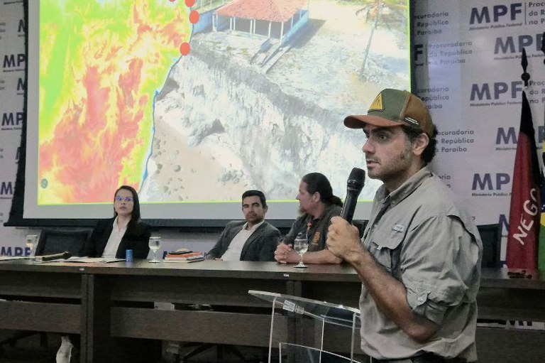 Foto mostra o pesquisador Rubson Maia, um homem branco de barba e boné, falando ao microfone durante o Painel Científico do Preamar. Ao fundo, vê-se a mesa que conduz os trabalhos, bem como um slide da apresentação.