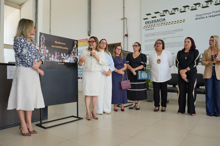 Um grupo de mulheres está em um ambiente institucional, diante de um painel com fotos e o título “Mulheres invisibilizadas”. Uma mulher fala ao microfone enquanto outra permanece ao lado do painel. Ao fundo, há uma placa grande com informações sobre a Delegacia Online da Paraíba.
