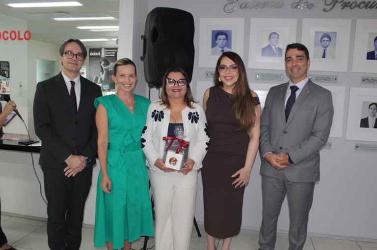 Cinco pessoas estão alinhadas em um ambiente institucional, vestidas de forma formal. A mulher ao centro segura um presente embrulhado com fita vermelha, sugerindo um momento de homenagem. Atrás delas há uma parede com retratos e a inscrição “Galeria dos Procuradores”, reforçando o contexto oficial. Todos posam para a foto com expressões neutras ou leves sorrisos.