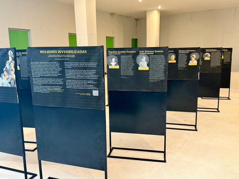 A imagem mostra uma exposição em ambiente interno, com painéis pretos alinhados em fileiras. Cada painel traz textos e retratos de mulheres brasileiras que tiveram suas histórias invisibilizadas ao longo do tempo. O título em destaque é “Mulheres Invisibilizadas”, e o espaço expositivo possui paredes brancas, portas verdes e colunas.
