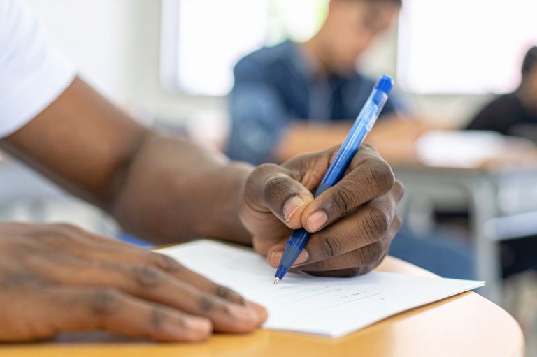 Close-up das mãos de uma pessoa negra escrevendo em uma folha de papel branca com uma caneta azul, durante uma prova ou aula. Ao fundo, de forma desfocada, aparecem outros estudantes em uma sala de aula iluminada.