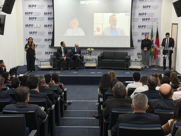 Auditório institucional lotado, com público sentado diante de um palco. Ao fundo, uma grande tela exibe duas pessoas por videoconferência. No palco, há autoridades sentadas, um homem falando ao púlpito e uma intérprete de Libras. As paredes exibem a identificação do MPF na Paraíba.
