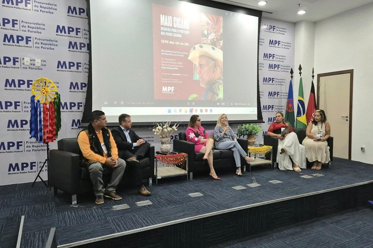 Em um auditório do Ministério Público Federal na Paraíba, seis pessoas estão sentadas em dois sofás pretos sobre um palco, participando de uma mesa de debate. Ao fundo, há uma tela de projeção exibindo um cartaz com o título “Maio Cigano – Desafios para a proteção aos povos ciganos”.