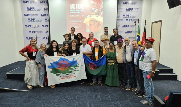 Um grupo de pessoas está reunido em um auditório, posando para uma foto coletiva. Ao fundo, há um painel com o texto MPF Procuradoria da República na Paraíba. Algumas pessoas seguram bandeiras da comunidade cigana, com fundo azul e verde e uma roda vermelha ao centro. Atrás do grupo, vê-se um cartaz anunciando o evento Maio Cigano Desafios para a proteção dos povos ciganos, realizado em João Pessoa, Paraíba