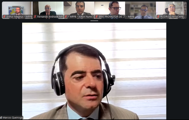 Captura de tela de reunião virtual, com seis participantes exibidos em miniaturas na parte superior. Em destaque, um homem de terno e gravata, usando fones de ouvido com microfone, aparece diante de uma janela com persianas claras.