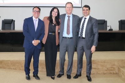 A imagem retrata quatro profissionais posando sorridentes em um plano médio, posicionados à frente da bancada de um auditório. O grupo é composto por três homens vestindo ternos e gravatas em tons de azul e cinza, e uma mulher vestindo uma calça preta com um blazer em tom terroso (caramelo). Eles estão alinhados lado a lado em uma postura amigável e formal, típica de registros de encerramento de eventos institucionais. O fundo mantém a coerência com as imagens anteriores, exibindo a estrutura de madeira escura da mesa de autoridades, poltronas de couro e o piso acarpetado em tom neutro, reforçando o ambiente de uma conferência jurídica ou administrativa.