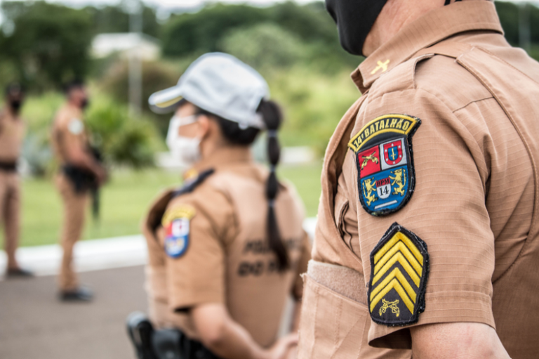 Plano médio em ângulo lateral de policiais militares brasileiros fardados em um ambiente externo. Em primeiro plano, o braço de um policial exibe o emblema do "14º Batalhão" e as divisas de graduação de Sargento. Ao fundo, outros policiais aparecem desfocados.