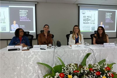 Foto de uma das mesas do evento com quatro mulheres sentadas e dois telões atrás