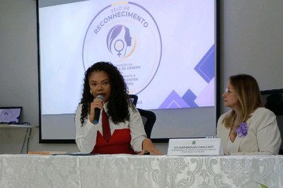 Foto mostra a servidora Maria Janaina falando ao microfone e atrás um telão do evento