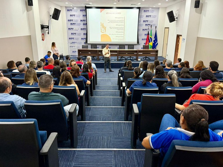 Em um auditório, pessoas estão sentadas em cadeiras azuis assistindo a uma palestra. No palco, um homem fala diante de uma tela projetada com o título “Mês das Mulheres”, enquanto uma mulher está de pé ao lado. Atrás deles há bandeiras e um painel com a sigla “MPF”.
