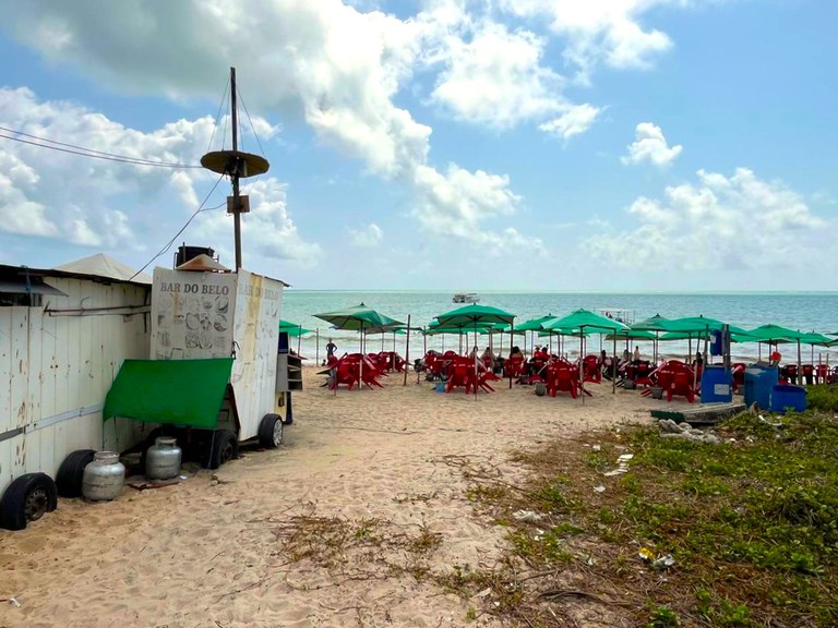 A imagem mostra uma área de praia com várias mesas e cadeiras vermelhas organizadas sob guarda-sóis verdes, ocupando uma parte significativa da faixa de areia. À esquerda, há uma estrutura rústica com placas que indicam Bar do Belo e botijões de gás próximos. O céu está azul com nuvens espalhadas, e o mar aparece ao fundo, tranquilo, com um barco visível no horizonte. No canto inferior direito, há vegetação rasteira e alguns resíduos no chão, indicando um espaço que mistura uso comercial com áreas naturais.