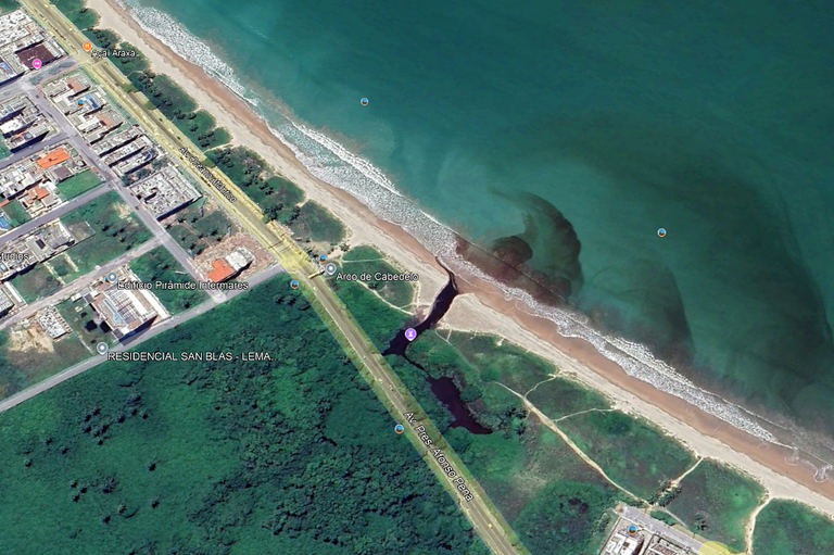 A imagem aérea mostra um trecho do litoral de Intermares, em Cabedelo (PB), com destaque para a foz do maceió do rio Jaguaribe, que deságua no mar. No centro, é visível o ponto onde o curso d¿água cruza a faixa de areia e forma uma mancha escura no oceano, indicativa do encontro entre as águas do rio e do mar. À esquerda, há áreas urbanizadas com prédios e ruas paralelas à orla; à direita e abaixo, aparecem áreas verdes de vegetação nativa.