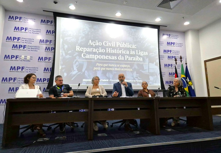 Em uma sala de conferências do Ministério Público Federal na Paraíba, seis pessoas estão sentadas lado a lado atrás de uma mesa comprida, participando de um evento oficial. Ao fundo, há um telão com o título Ação Civil Pública: Reparação Histórica às Ligas Camponesas da Paraíba”e o lema Para que nunca se esqueça, para que nunca mais aconteça.