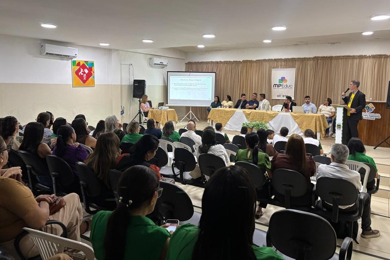 Descrição da imagem: Fotografia em plano geral e ângulo visto de trás mostra um auditório amplo durante uma audiência pública do projeto MPEduc, em Cruz do Espírito Santo (PB). Em primeiro plano, dezenas de pessoas estão sentadas de costas em fileiras de cadeiras pretas, acompanhando o evento; algumas vestem blusas verdes. Ao fundo, no palco, há uma mesa comprida coberta com toalhas branca e dourada, onde estão sentadas cerca de sete pessoas. À esquerda da mesa, uma projeção em um telão exibe um texto com o título "Escola em Tempo Integral". À direita, um homem de terno preto fala ao microfone em pé atrás de um púlpito com o logotipo da prefeitura local. Atrás da mesa, há uma cortina de tecido bege e um banner vertical com a logomarca colorida do "MPEduc". O ambiente é bem iluminado por luminárias embutidas no teto branco e possui aparelhos de ar-condicionado instalados nas paredes.
