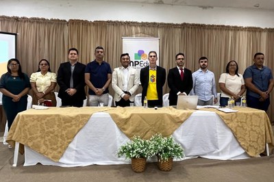 Descrição da imagem: Fotografia em plano médio e visão frontal mostra um grupo de dez pessoas posando de pé, lado a lado, atrás de uma mesa comprida durante o evento do MPEduc. O grupo é composto por seis homens e quatro mulheres, de aparências e vestimentas variadas que incluem ternos, camisas sociais e blusas casuais. Ao centro, um dos homens destaca-se vestindo uma camiseta amarela sob um paletó preto. A mesa à frente deles está decorada com uma toalha branca e detalhes em tecido dourado com arabescos, tendo ao chão dois vasos com pequenas flores brancas e folhagens verdes. Ao fundo, uma cortina de tecido bege cobre toda a extensão da parede, e um banner vertical com o logotipo colorido do "MPEduc" está posicionado centralizado atrás do grupo.