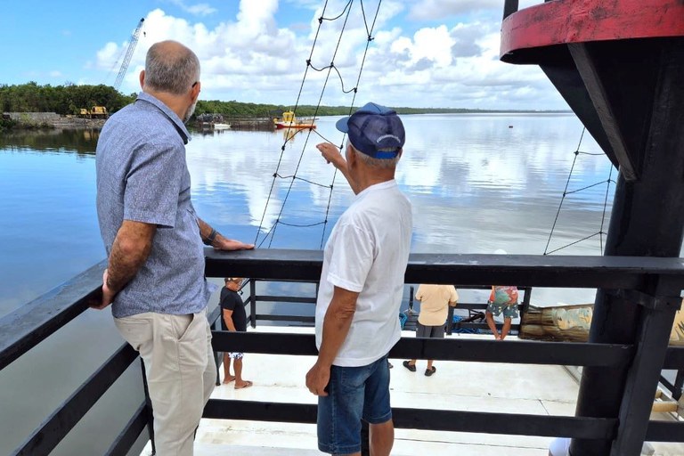 Dois homens estão de pé sobre a plataforma de um barco, observando o estuário do rio Paraíba. Um deles aponta para a margem, onde há máquinas e obras em andamento. Ao fundo, o céu está azul com nuvens esparsas e a água reflete a paisagem ao redor.