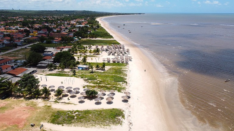A imagem mostra uma praia com areia clara e águas escuras. Há várias barracas de palha espalhadas, algumas palmeiras e uma vegetação ao redor. Pode-se visualizar degradação da faixa de vegetação de restinga e construções de imóveis sobre esta.
